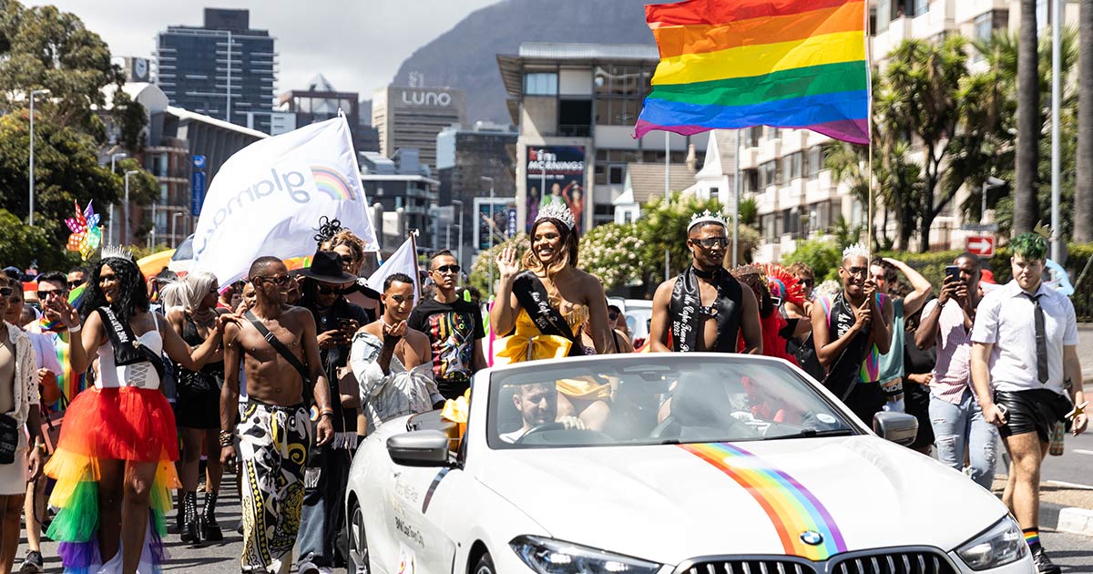 A photo of the colourful parade being led by Mr and Miss Cape Town Pride sitting in a convertible vehicle.
