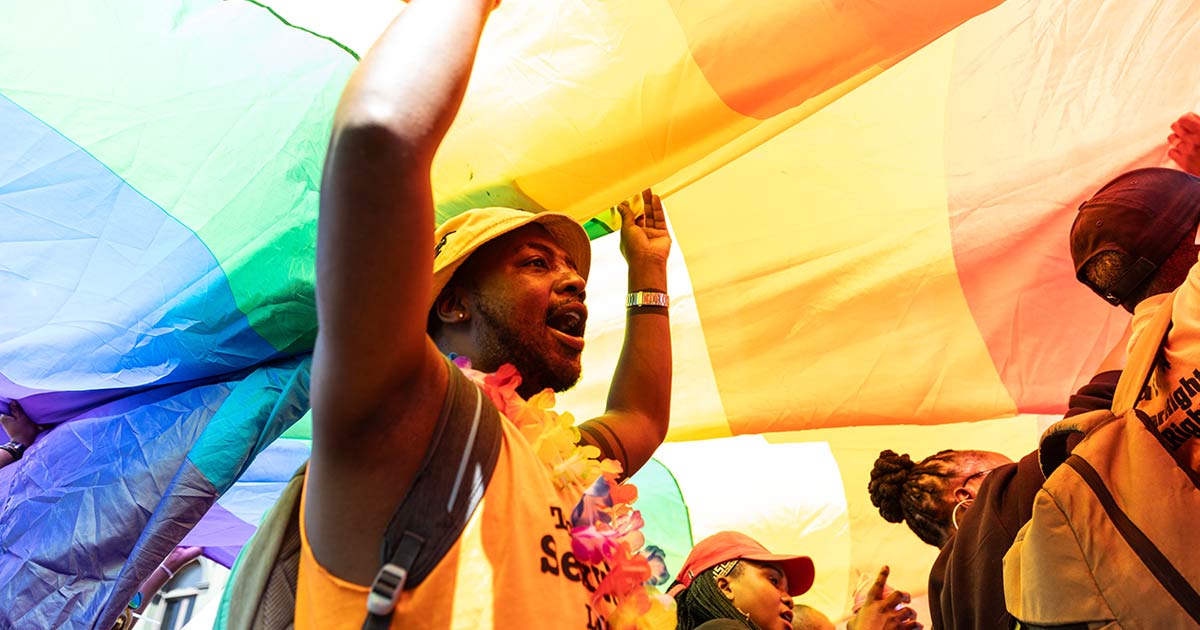 A photo of a joyful man holding up a huge Pride rainbow flag.