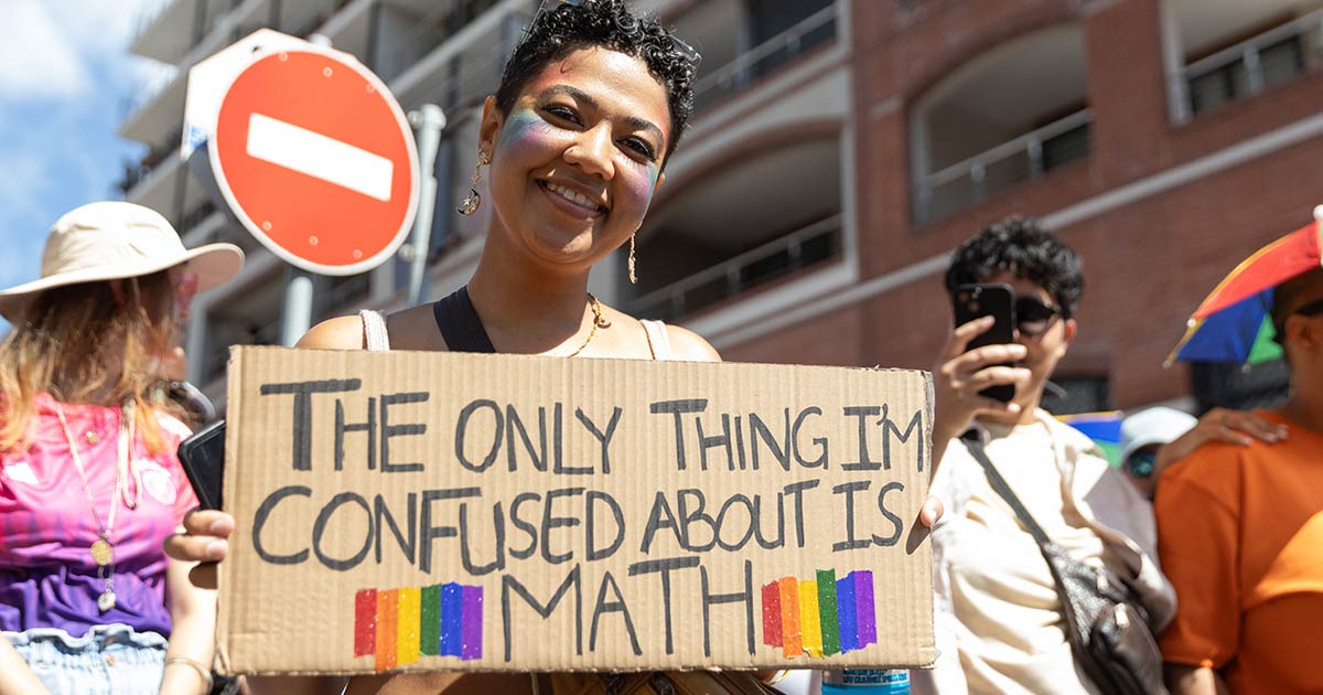 A photo of a young individual, Keshia Jansen, smiling in the Cape Town Pride parade while holding up a placard that reads: "The only thing I'm confused about is math."